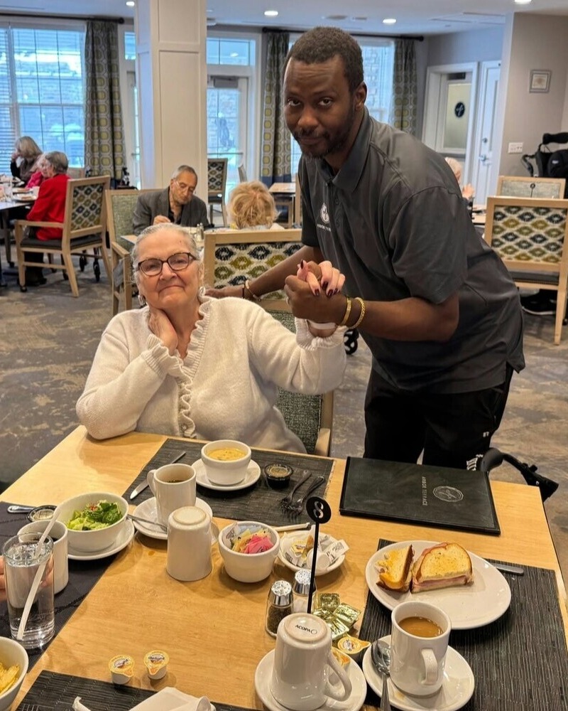 A smiling woman in a white cardigan holds hands with a man in a dark shirt who leans in beside her at a dining table, sharing a warm moment in a bright dining room at senior living with on-site dining in Exton, PA, a friendly place for senior living near Paoli, PA.