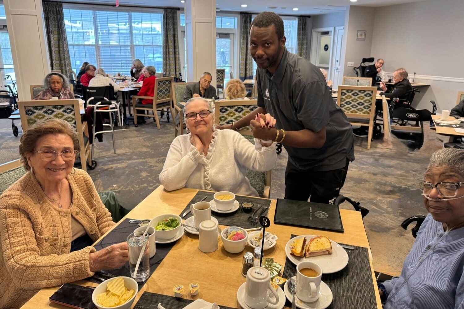 Residents enjoy a warm lunch together in a bright dining room while a staff member lends a helping hand, reflecting the friendly atmosphere of senior living with on-site dining in Exton, PA and active senior communities in Exton, PA.