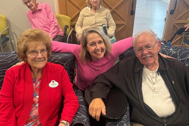 Three residents share smiles and warm hugs in a cozy lounge, enjoying each other's company, a heartfelt moment reflecting senior housing near Malvern, PA and resort-style senior living Exton, PA.