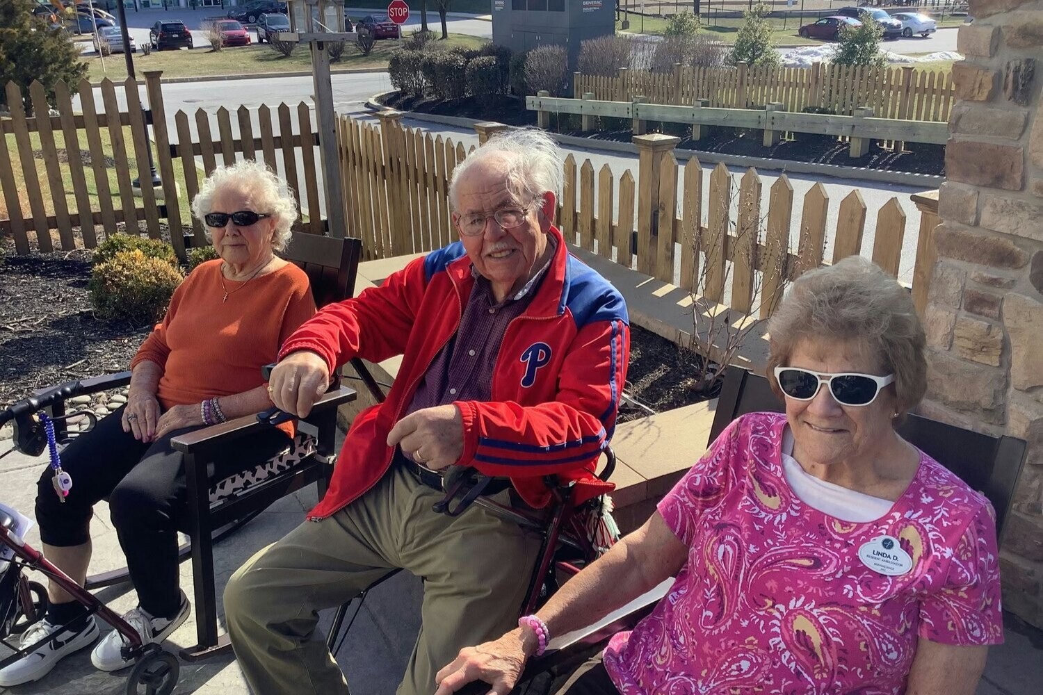 Three residents enjoy a sunny afternoon on the porch, surrounded by a picket fence garden, embodying the relaxed charm of senior living near hiking trails in Chester County, PA and senior living in Malvern, PA.