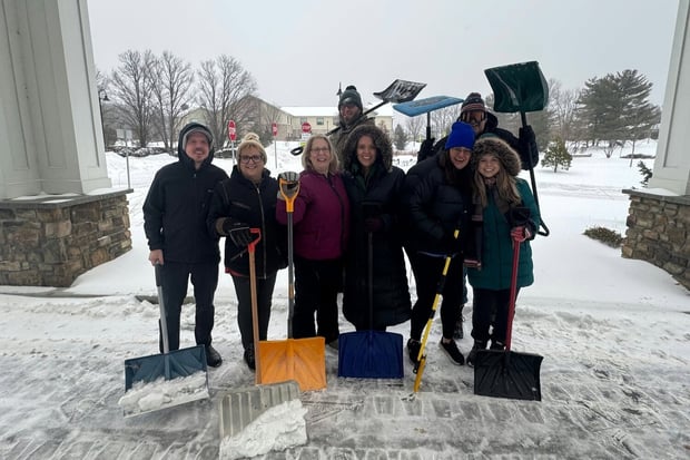 A cheerful team pauses with snow shovels in hand after clearing a snowy entrance, showing the dedication behind senior living in Exton, PA and retirement communities near Downingtown, PA.
