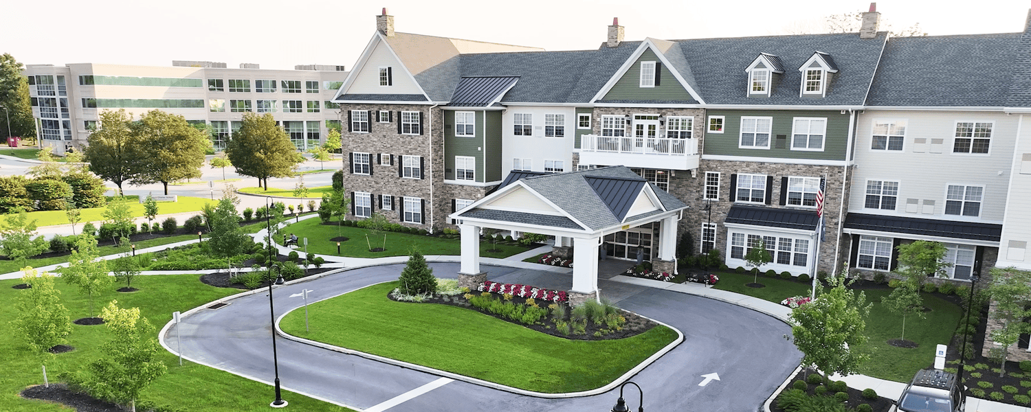 An aerial view of Arbor Terrace Exton, with a stone and siding exterior, dormer windows, a covered front entry, and a circular drive around a lush lawn and flower bed, one of the inviting memory care communities in Exton, PA, and personal care communities near Paoli, PA.