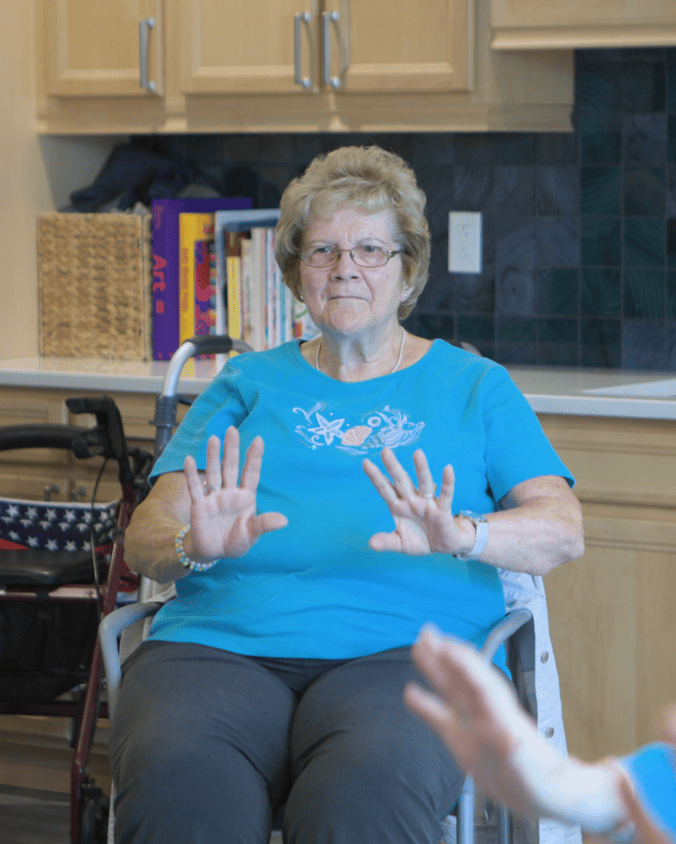 A cheerful woman in a turquoise tee and glasses raises both hands with a bright smile during an activity in a cozy room with wood cabinets and shelves of books, capturing the fun of senior living in King of Prussia, PA, and senior living near South Coventry, PA.
