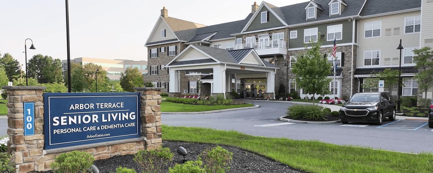 The welcoming entrance of Arbor Terrace Exton, with a stone monument sign, a covered front entry, and a stone and gray siding exterior framed by trees and manicured landscaping, offering personal care in Exton, PA, and memory care near Exton, PA.