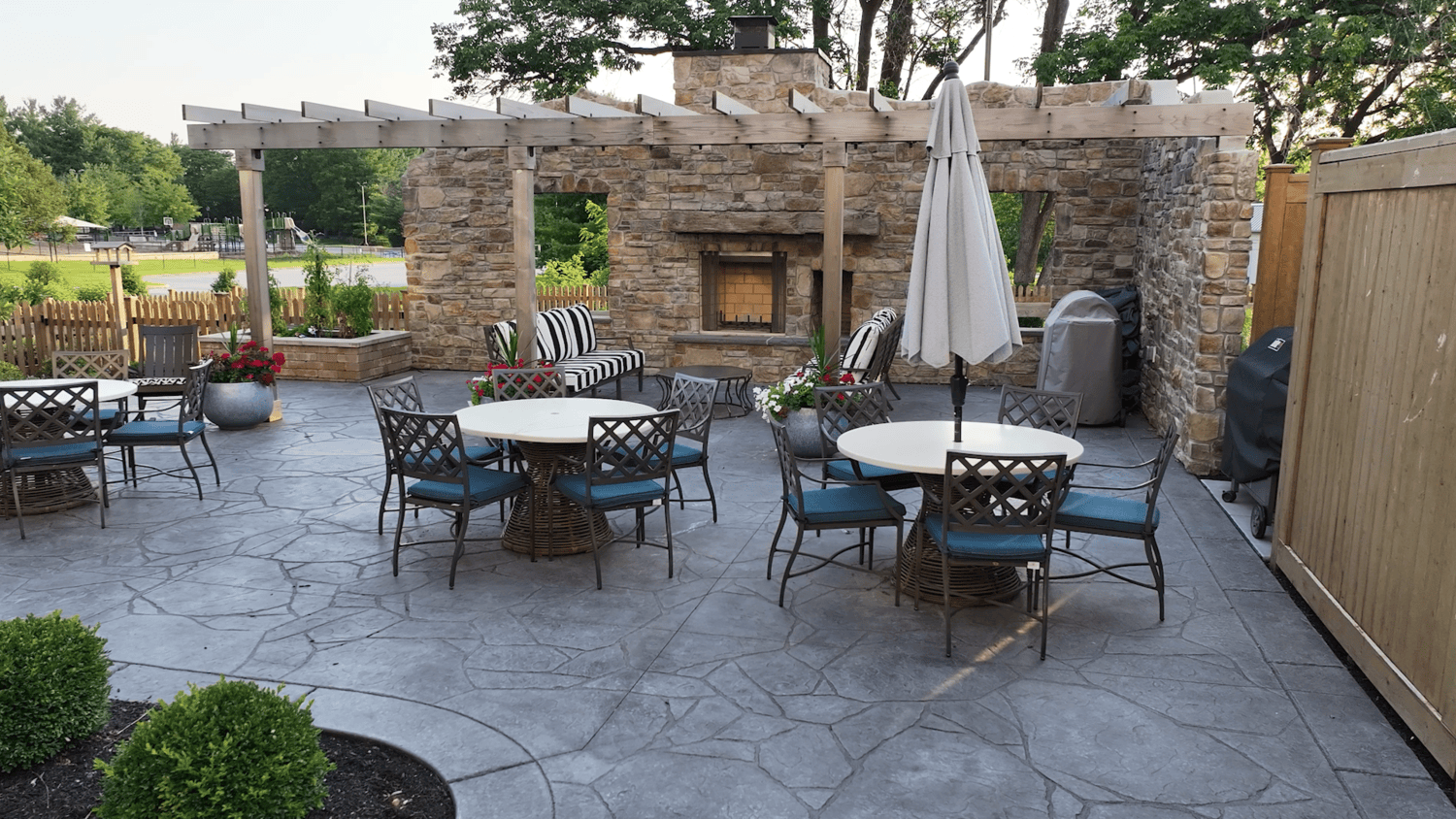 A stone patio with a pergola-covered outdoor fireplace, round tables with blue-cushioned chairs, a patio umbrella, and striped lounge seating framed by leafy trees, an inviting retreat for resort-style senior living Exton, PA, and assisted living in Malvern, PA.