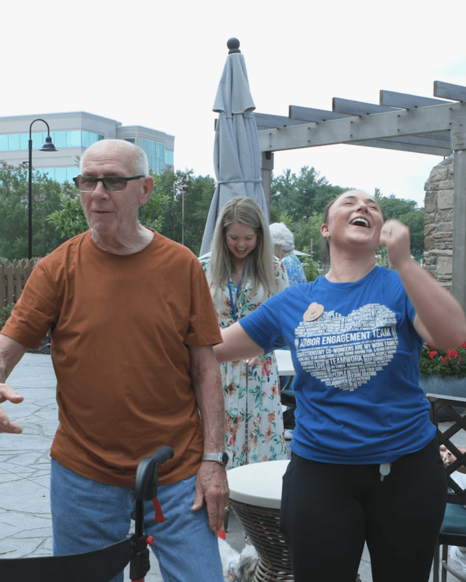 A man in an orange tee and a woman in a blue tee share a hearty laugh on a sunny patio with a wooden pergola and umbrella, showing the upbeat spirit of active senior living near Downingtown, PA, and a lively community for senior living in Coatesville, PA.