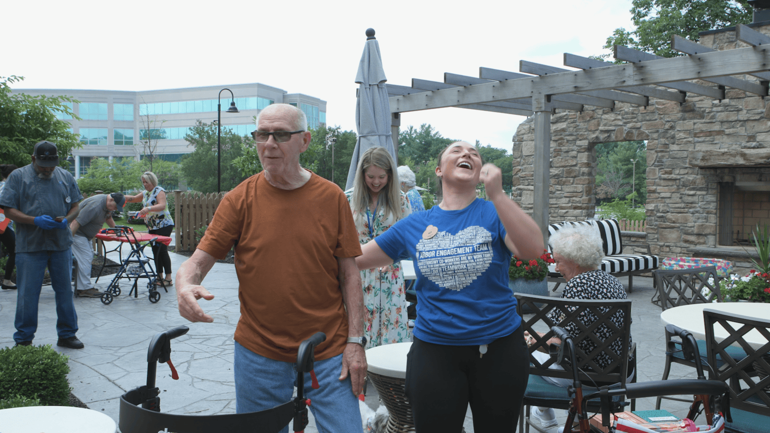 A man in an orange tee and a woman in a blue tee share a hearty laugh on a sunny patio with a wooden pergola and umbrella, showing the upbeat spirit of active senior living near Downingtown, PA, and a lively community for senior living in Coatesville, PA.