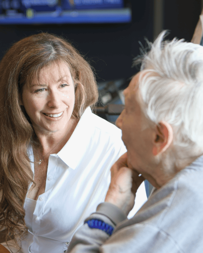 A woman in a white blouse smiles warmly while chatting with a person with white hair who rests their chin on one hand, sharing an easy conversation that reflects senior living near Paoli, PA, and the friendly connections found in senior living in Malvern, PA.