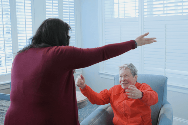A smiling woman in a bright orange jacket sits in a blue armchair and stretches her arms wide, mirroring a woman in a maroon top during a lively moment in a sunny room with white shutters, a joyful scene at senior living in Exton, PA, and senior living near South Coventry, PA.