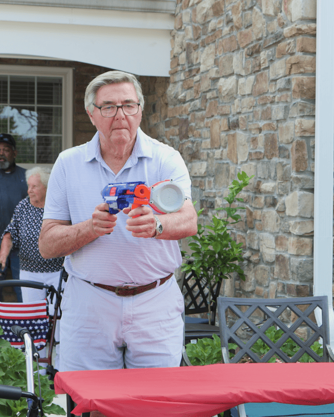 A smiling man in a light blue polo holds a festive red, white, and blue item and a cup at a lively outdoor celebration beside a stone building, a joyful moment at senior living in Exton, PA, and a welcoming option for senior living in King of Prussia, PA.