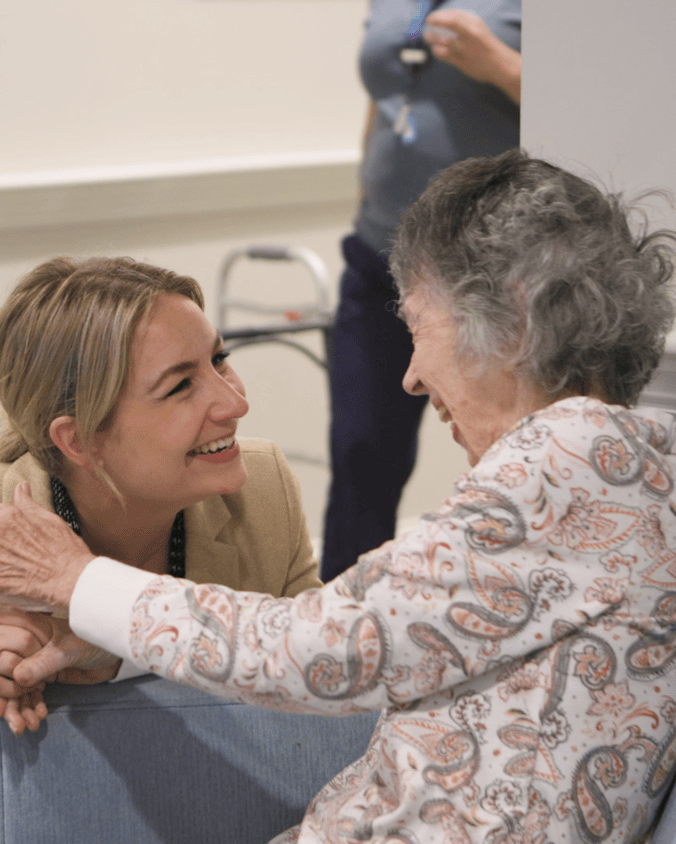 A smiling woman with blonde hair leans in close to a gray-haired woman in a paisley top, their hands clasped as they share a joyful moment, a heartwarming scene at senior living in Exton, PA, and a caring community for senior living near Phoenixville, PA.