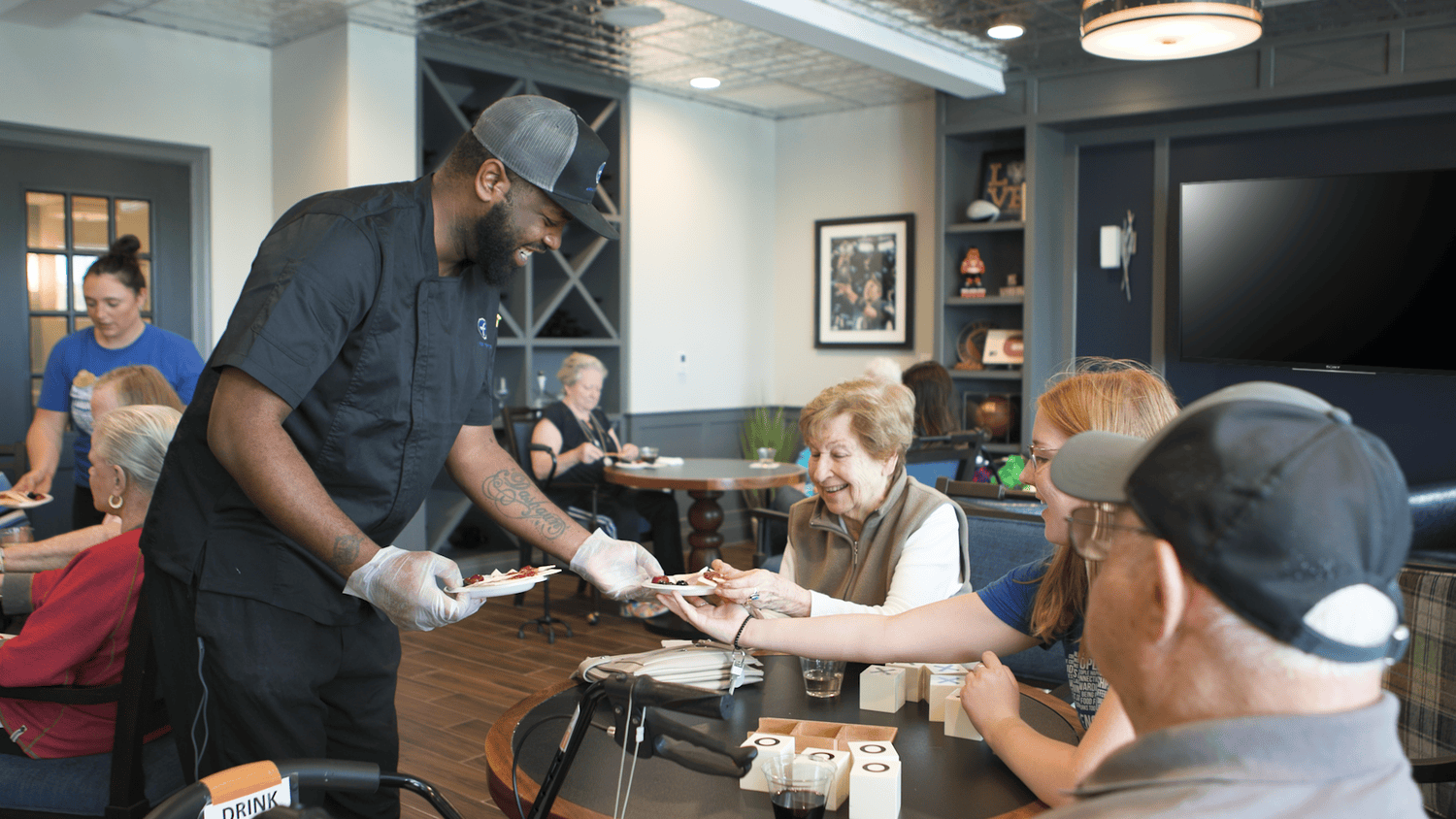 A man in a chef coat and cap serves a plate to a smiling woman at a table while others gather nearby in a cozy lounge with framed art and dark shelving, a friendly moment at senior living in Coatesville, PA, and a sociable spirit found in senior living near Paoli, PA.