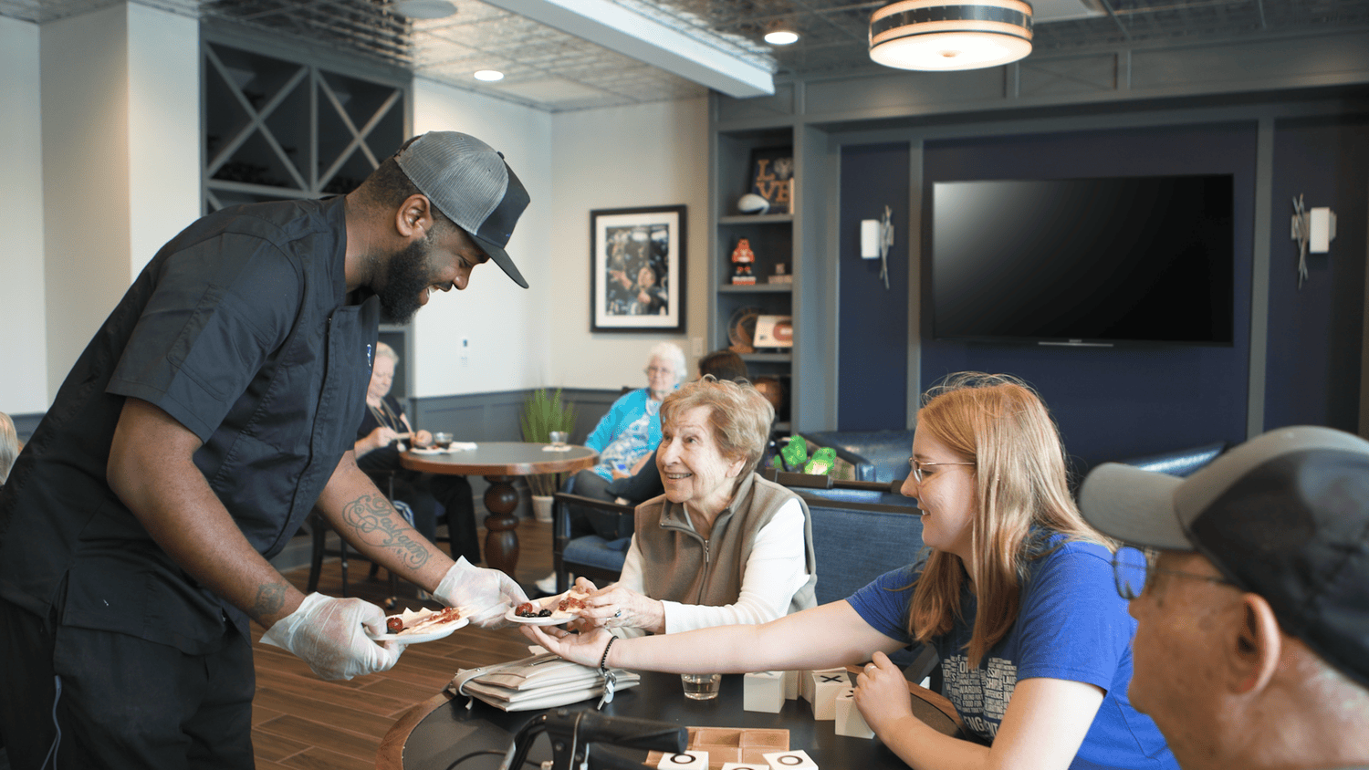A culinary staff member serves residents and their guests in a warmly decorated dining lounge at Arbor Terrace Waugh Chapel, capturing the attentive, resort-style dining experience and welcoming community atmosphere at this senior living community in Gambrills, MD.
