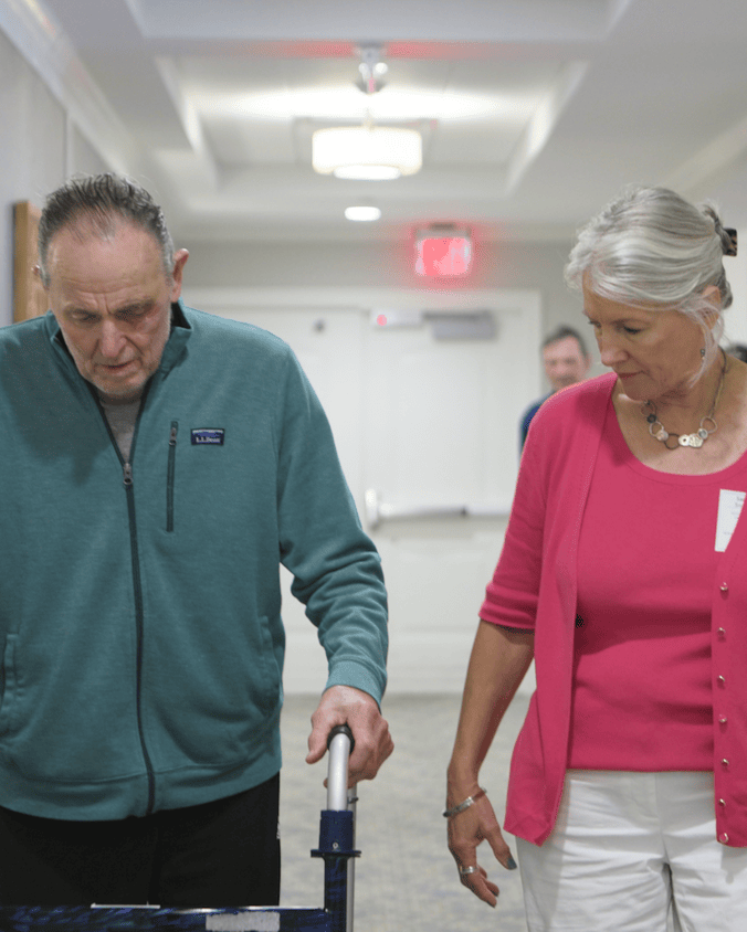 A man in a teal jacket and a woman in a pink cardigan walk side by side down a bright hallway, sharing an easy stroll together that reflects the warm, supportive setting of senior living near West Chester, PA, and senior living in Downingtown, PA.