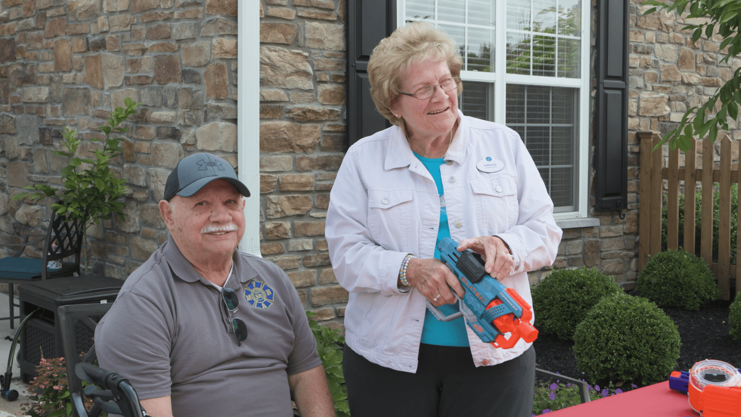 A smiling man in a gray polo and ball cap sits beside a laughing woman in a white jacket holding a colorful toy blaster outside a stone building with trimmed shrubs, a fun outdoor moment at senior living in Malvern, PA, and senior living in Coatesville, PA.