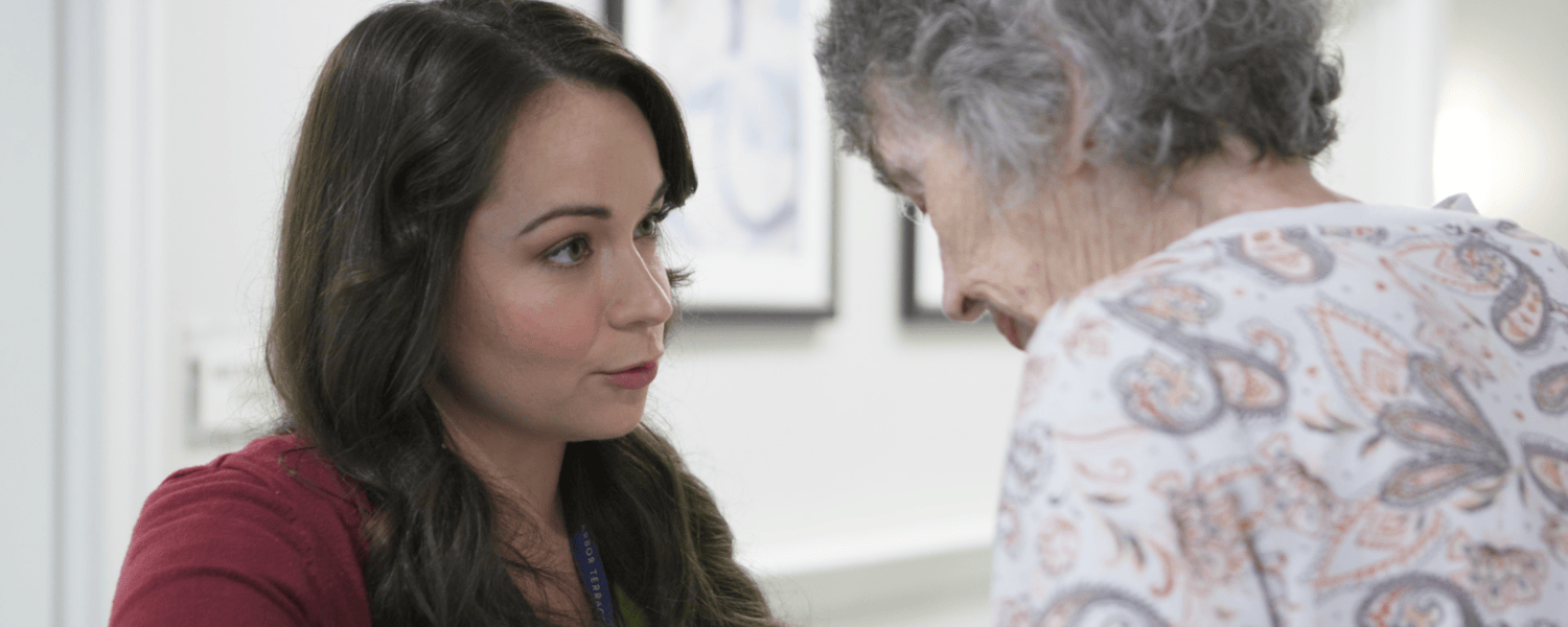 A woman with long dark hair in a red cardigan leans in face to face with a gray-haired woman in a paisley top, sharing a quiet, attentive moment in a bright hallway, a gentle scene of connection at senior living near Phoenixville, PA, and senior living in Malvern, PA.