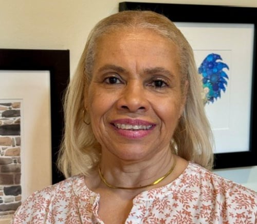 Ana DaSilva, Memory Care Engagement Program Manager, smiles in a floral top and gold necklace in front of framed art, enriching memory care near Exton, PA, and senior living in King of Prussia, PA.