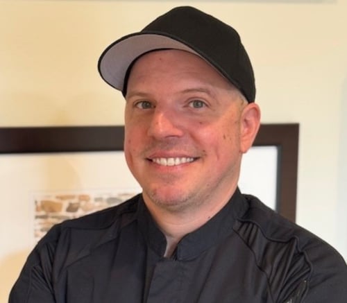 Jared Mininger, Dining Director, smiles in a black ball cap and dark chef coat in front of framed art, leading senior living with on-site dining in Exton, PA, and senior living in Coatesville, PA.