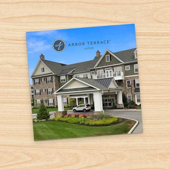 Square photo of Arbor Terrace Exton multi-story building exterior with white columned entrance, landscaped flower beds, and parked car under portico, displayed on a printed card against a wood-grain background.