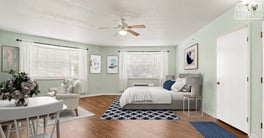 A spacious, light-filled studio apartment at Barrington Terrace of Fort Myers, FL, featuring hardwood floors, sage green walls, a ceiling fan, large windows, a cozy sitting area, and a neatly styl