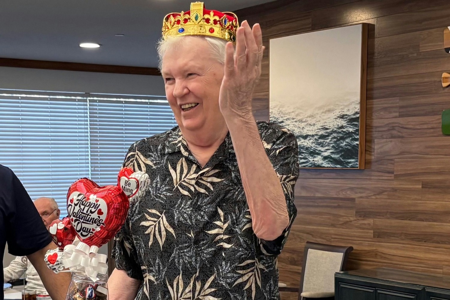 A resident wearing a gold crown and waving with a big smile, celebrating Valentine's Day at Barrington Terrace, a senior living community in Fort Myers, FL, with festive heart-shaped balloons nearby.
