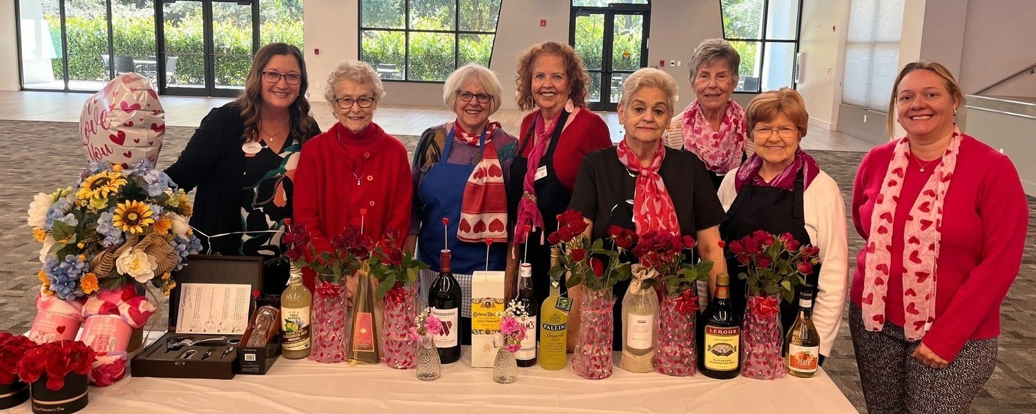A cheerful group poses behind a festively decorated table with flowers, wine bottles, and gifts at a Valentine's Day celebration at Barrington Terrace of Fort Myers, a senior living community in Fort Myers, FL. 