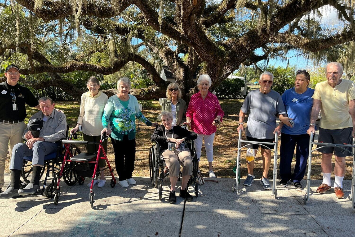 A group of residents and staff enjoy a sunny outdoor outing beneath a large oak tree at this senior living community in Fort Myers, FL.