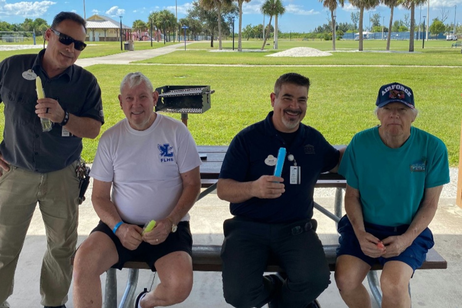 Residents relaxing and smiling together outdoors under a shaded area, with lush green lawns and palm trees in the background, enjoying the sunny Florida weather at Barrington Terrace in Fort Myers, FL.