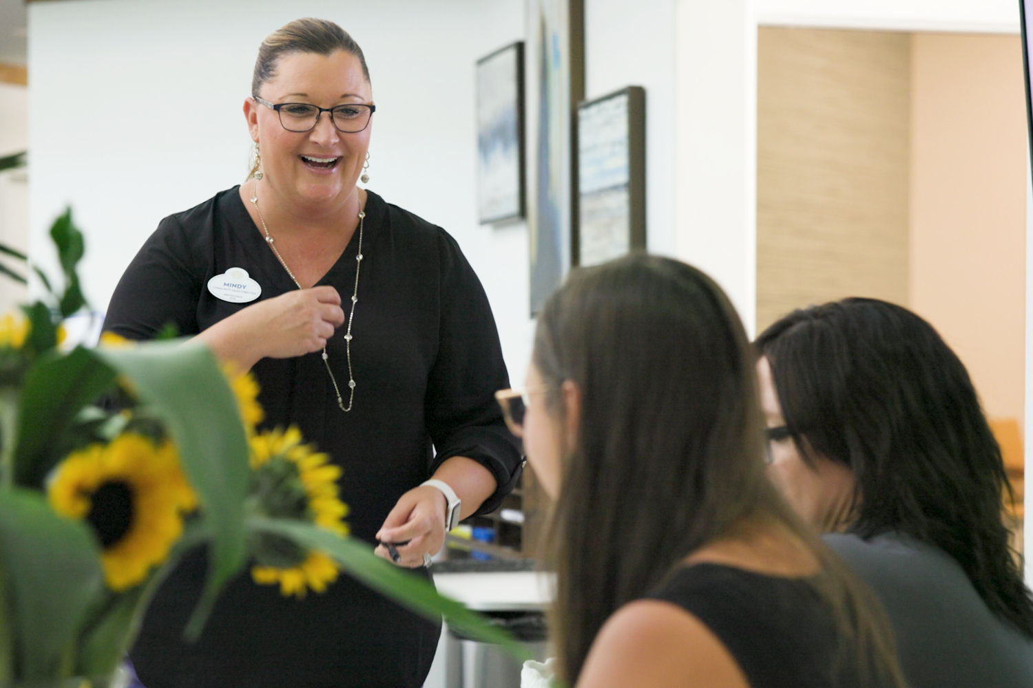 A smiling staff member holds sunflowers while chatting with residents in a bright common area at Barrington Terrace senior living in Fort Myers, FL.