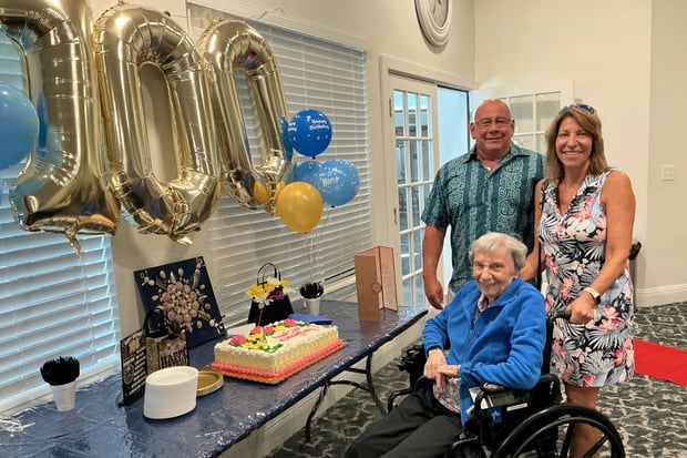 A resident in a wheelchair celebrates a milestone birthday with two companions beside gold balloon decorations at Barrington Terrace senior living in Fort Myers, FL.