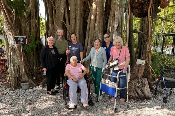 A cheerful group of residents and staff pose together beneath a large banyan tree during a community outing from Barrington Terrace senior living in Fort Myers, FL.