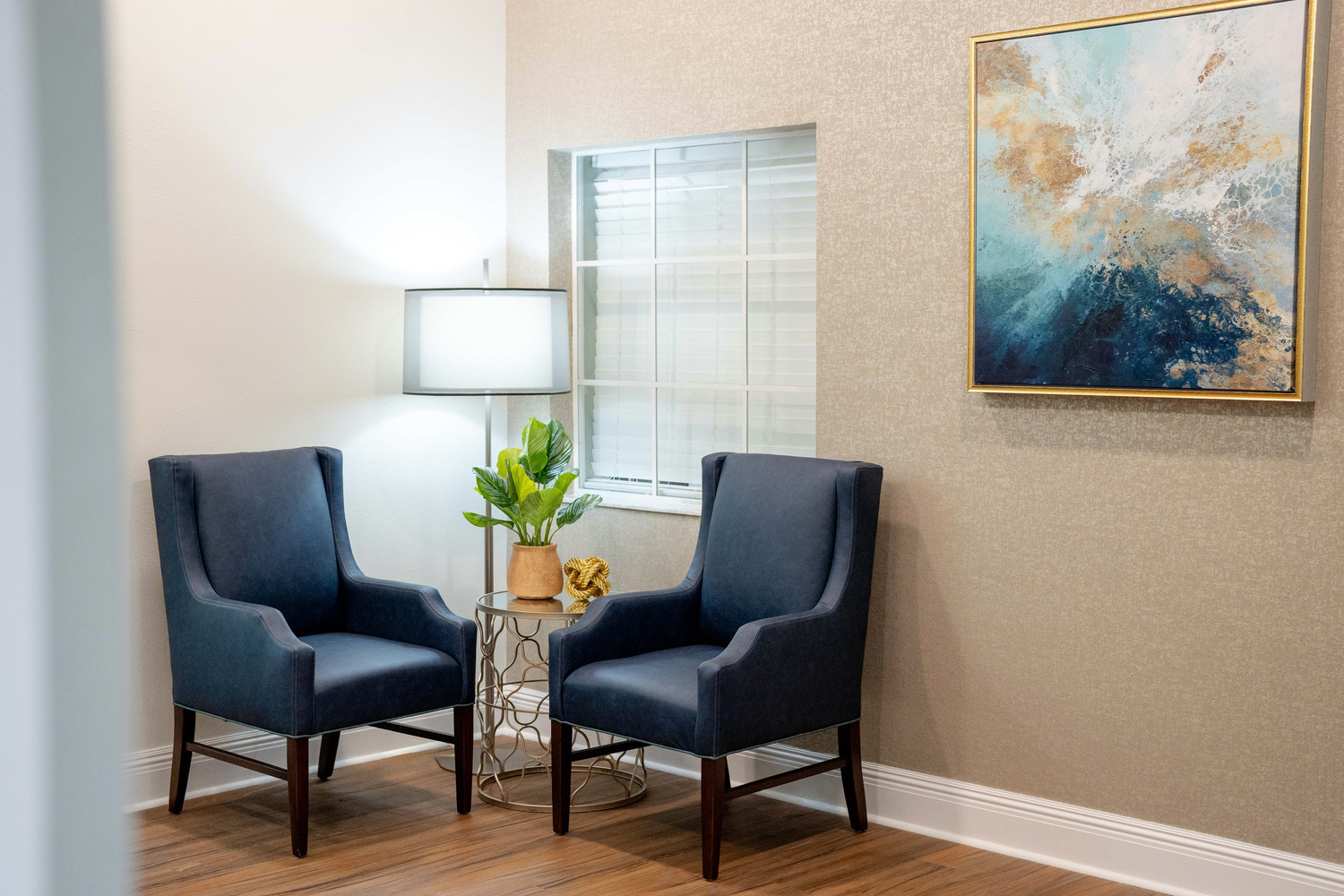 Cozy apartment corner with a navy armchair, soft lamp lighting, and a coastal-themed painting at this senior living community in Fort Myers, FL.