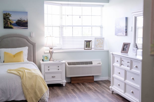 Sunlit apartment bedroom with white furniture, hardwood floors, and cheerful yellow bedding at Barrington Terrace senior living in Fort Myers, FL.