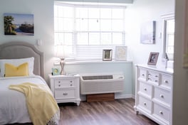 Sunlit apartment bedroom with white furniture, hardwood floors, and cheerful yellow bedding at Barrington Terrace senior living in Fort Myers, FL.