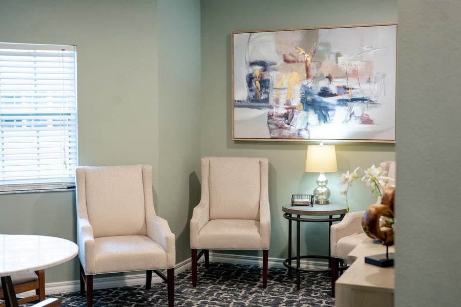 A bright, welcoming sitting area at assisted living in Fort Myers, FL, featuring cream wingback chairs, a round side table with a glowing lamp, fresh florals, and colorful abstract artwork on the wall.