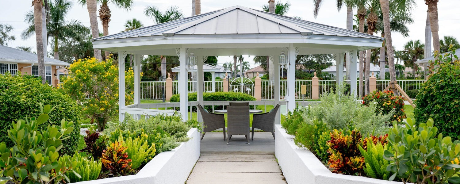 A white gazebo surrounded by lush tropical gardens and colorful plants offers a peaceful outdoor seating area at Barrington Terrace of Fort Myers, a senior living community in Fort Myers, FL.