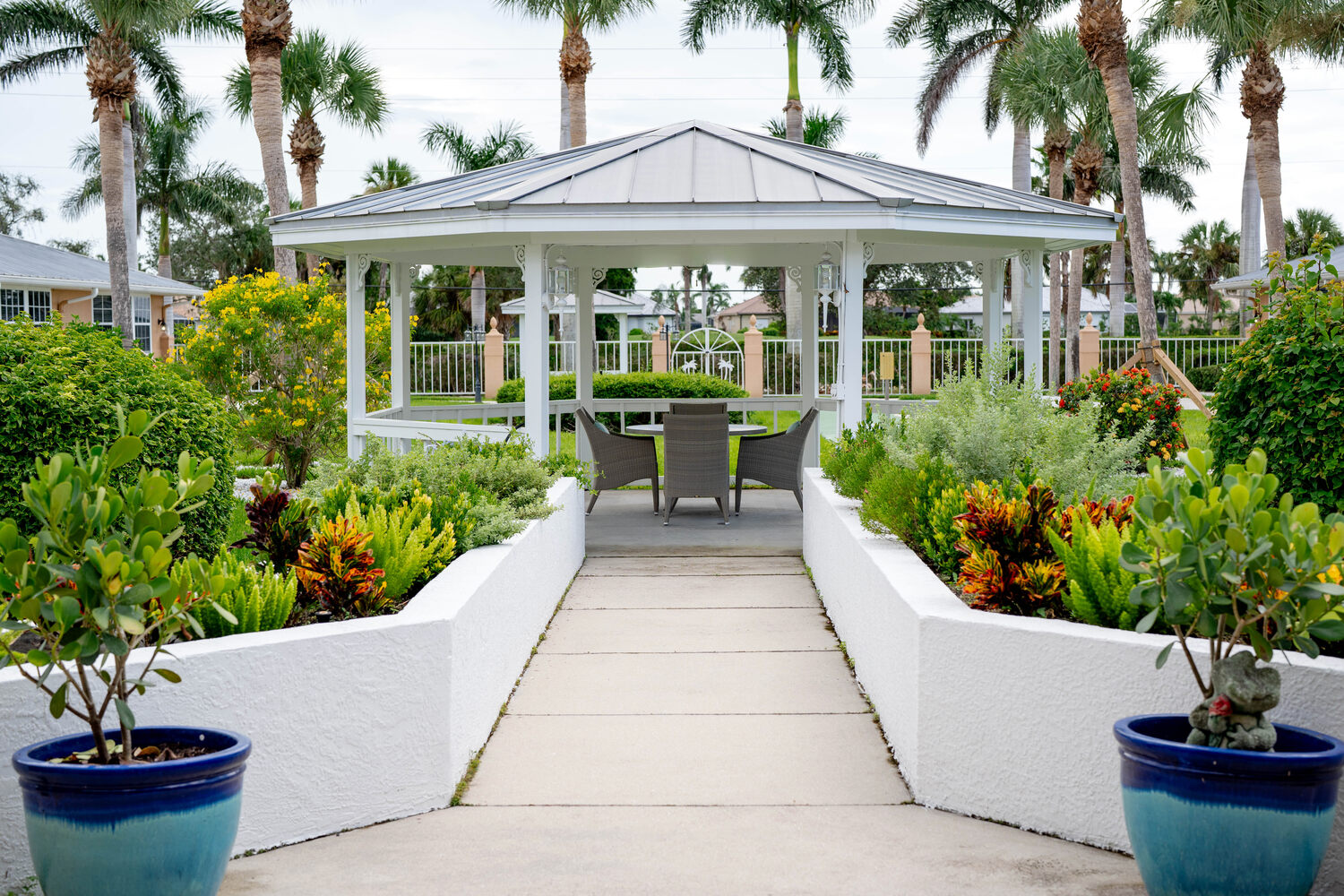 Scenic walkway leading to a white gazebo surrounded by vibrant tropical plants and palm trees at this senior living community in Fort Myers, FL.