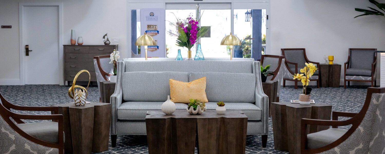 A welcoming seating area at Barrington Terrace, a senior living community in Fort Myers, FL, featuring a plush gray sofa, warm wood accent chairs, gold lamps, and fresh floral arrangements on a patterned area rug.