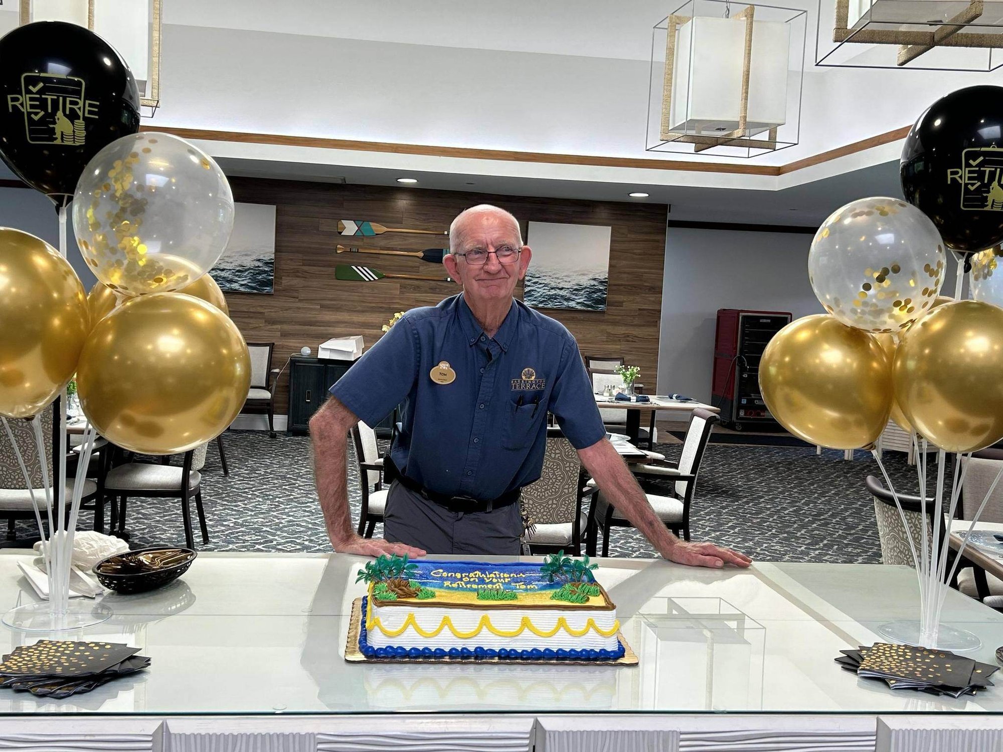 A staff member stands beside a decorated celebration cake surrounded by gold and black balloons in the dining room at Barrington Terrace senior living in Fort Myers, FL.