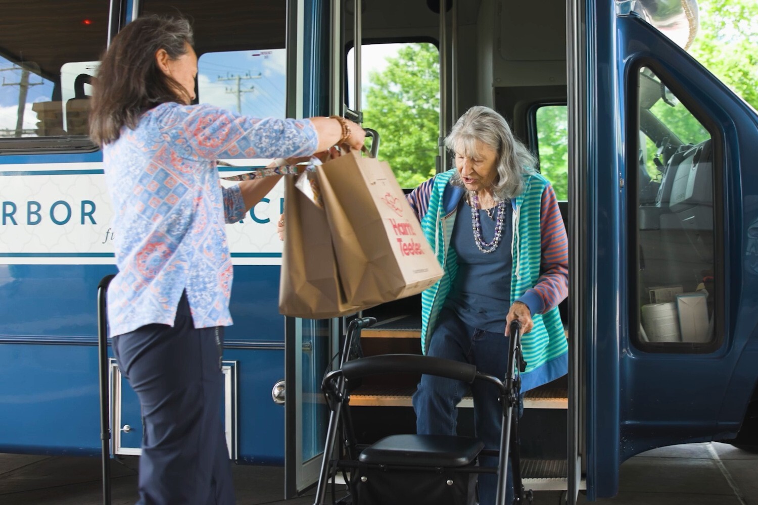 A senior woman with long silver hair and a striped hoodie steps off a shuttle bus as a woman in a paisley blouse hands her grocery bags, a helpful errand day that reflects the everyday ease of active senior communities in Atlanta, GA and top-rated senior living in Atlanta, GA.