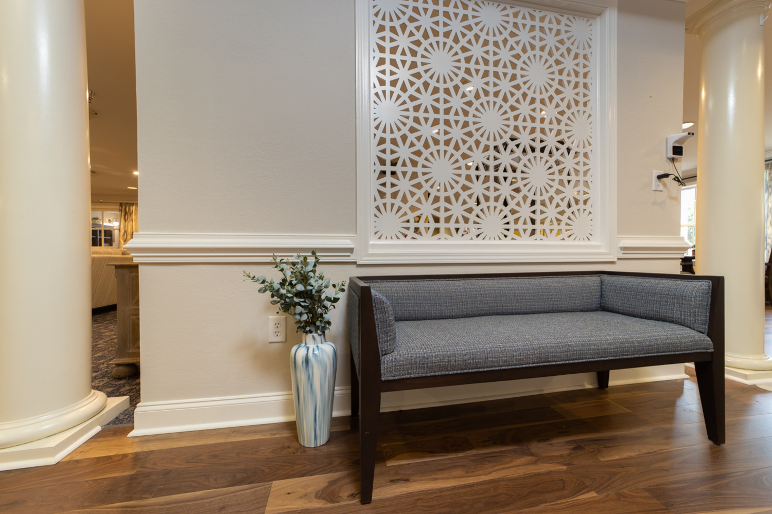 A bright common area features an elegant white lattice partition, a tufted gray bench, and a tall vase of greenery, showcasing the welcoming, boutique senior living Greenville offers within this senior living Greenville SC community.