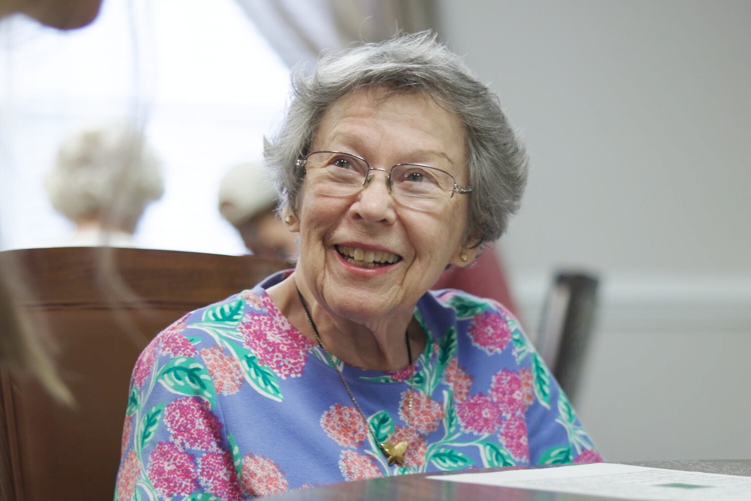 A resident smiles warmly in a close-up portrait, wearing a floral top and a delicate pendant necklace, radiating the joyful spirit and vibrant senior living lifestyle found throughout this senior living community.