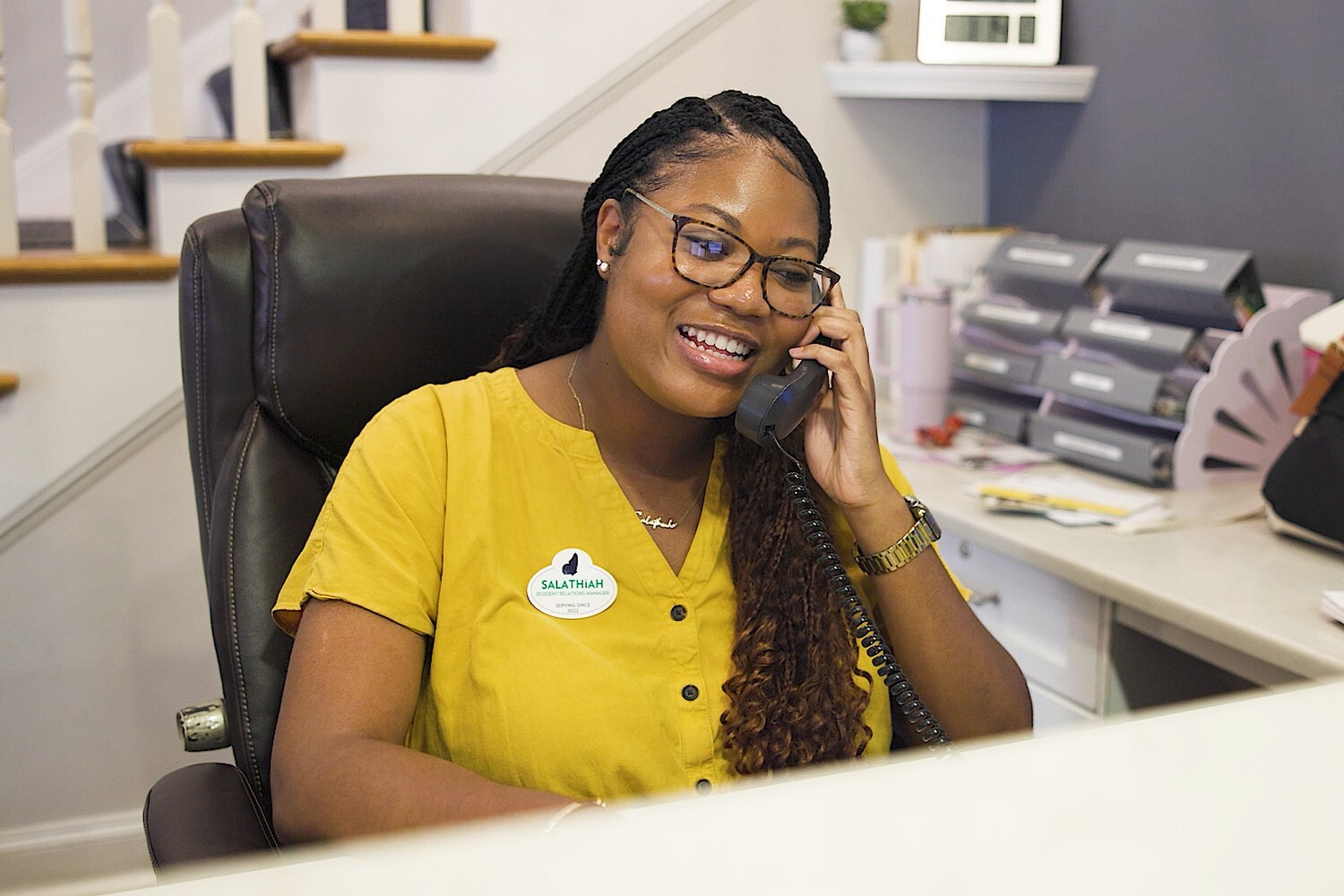A smiling team member answers the phone warmly at the front desk, wearing an Arbor Company polo, reflecting the friendly, welcoming spirit of a senior living community and its dedicated senior living team.