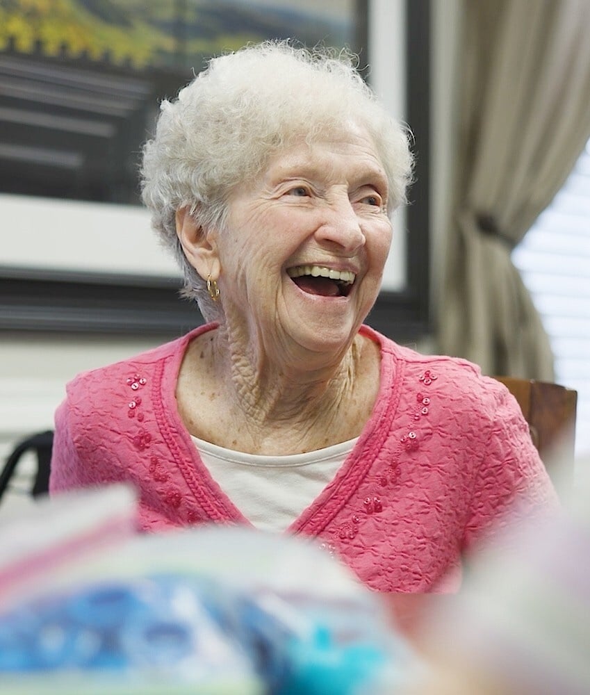 A resident laughs joyfully while opening colorful gifts, radiating warmth during a heartfelt celebration that reflects the vibrant senior living lifestyle found throughout this senior living community.