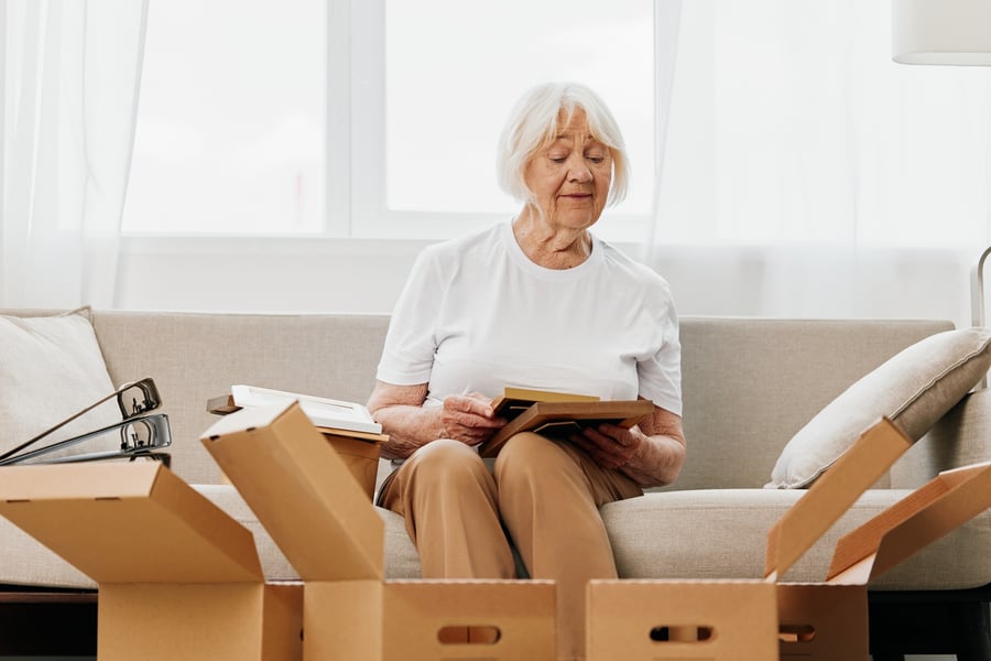 An older woman with white hair sits on a couch surrounded by open cardboard moving boxes, smiling as she looks through a photo album.