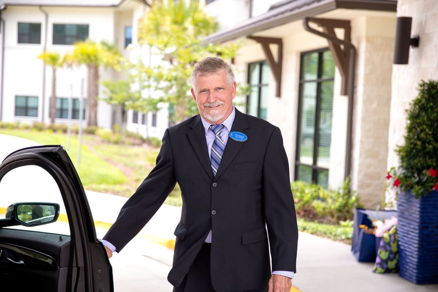 A staff member in a suit holds open a car door outside a senior living community, offering a warm and attentive welcome to residents and visiting families.