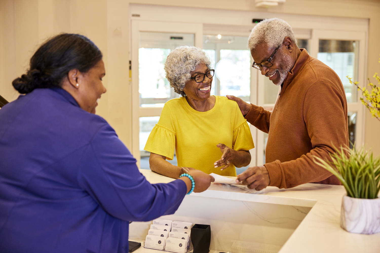 A staff member assists a resident couple with paperwork at the front desk, reflecting the attentive senior living in Sandy Springs, GA and Perimeter, GA.