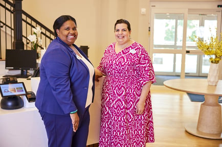 Two smiling team members stand together at the welcoming front desk of an assisted living community in Athens, GA, with a bright, airy lobby and fresh florals visible behind them.