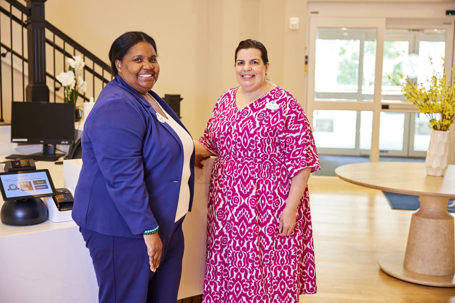 Two smiling team members stand together at the welcoming front desk of an assisted living community in Athens, GA, with a bright, airy lobby and fresh florals visible behind them.