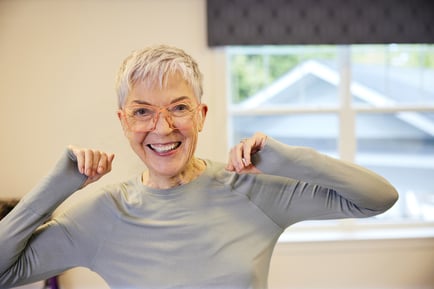 A smiling woman with short white hair and glasses stretches her arms in a bright, sunny room, reflecting the active and wellness-focused senior lifestyle at Arbor Terrace Herndon in Herndon, VA.